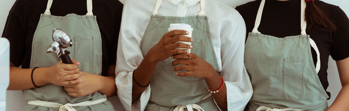 Three hospitality staff side by side in sage-green bib aprons over black and white tops, one holding an espresso portafilter and one a takeaway cup, framed across the chest and waist
