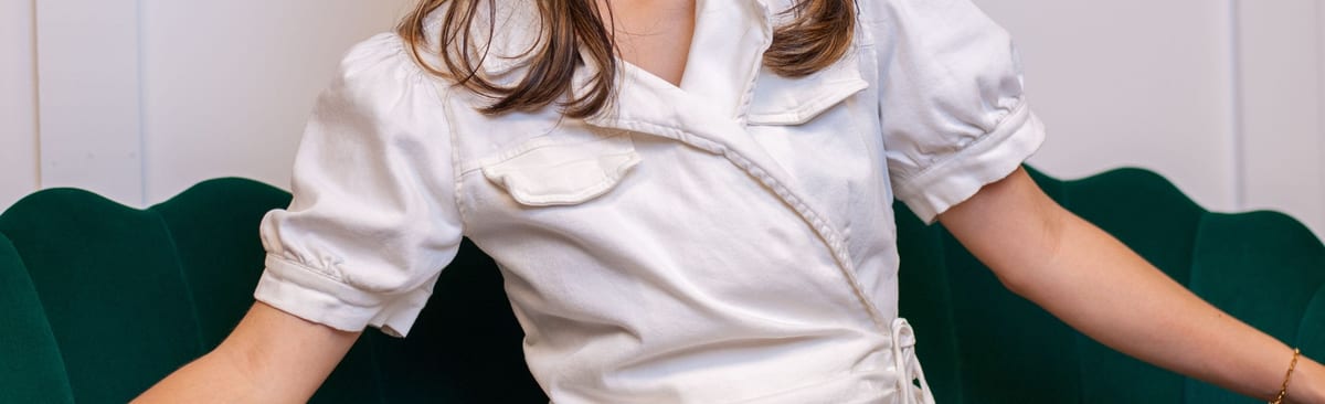 A white wrap-front dress with a buttoned chest pocket and short puff sleeves, worn against a dark green velvet sofa