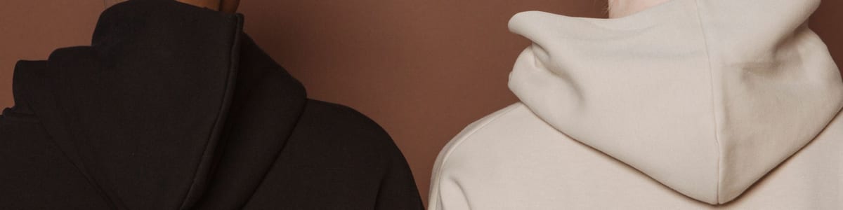 The back of a cream hoodie and a black hoodie, showing the hood seam and the upper back panel where a stitched logo sits
