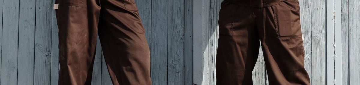 Brown work overalls and work trousers worn against a weatherboard wall, showing the hip and thigh pockets