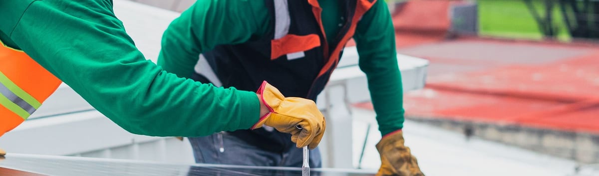 A worker in an orange hi-vis vest and hard hat fastening a rooftop solar panel with a hand tool, wearing heavy tan work gloves