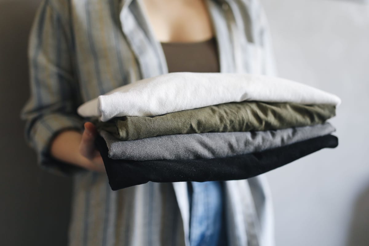 A stack of four plain folded tees, white, olive, grey and black, held out at chest height