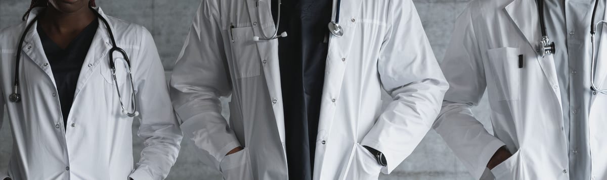 White lab coats worn open over dark V-neck tunics, stethoscopes at the neck, in a row against a grey concrete wall
